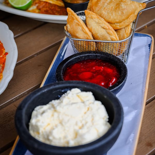 Fried empanadas in a metal basket with red and white dipping sauces on a wooden table.