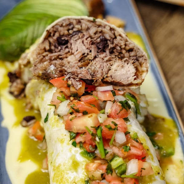 A large burrito, cut open to show shredded meat and rice, topped with pico de gallo and green sauce.