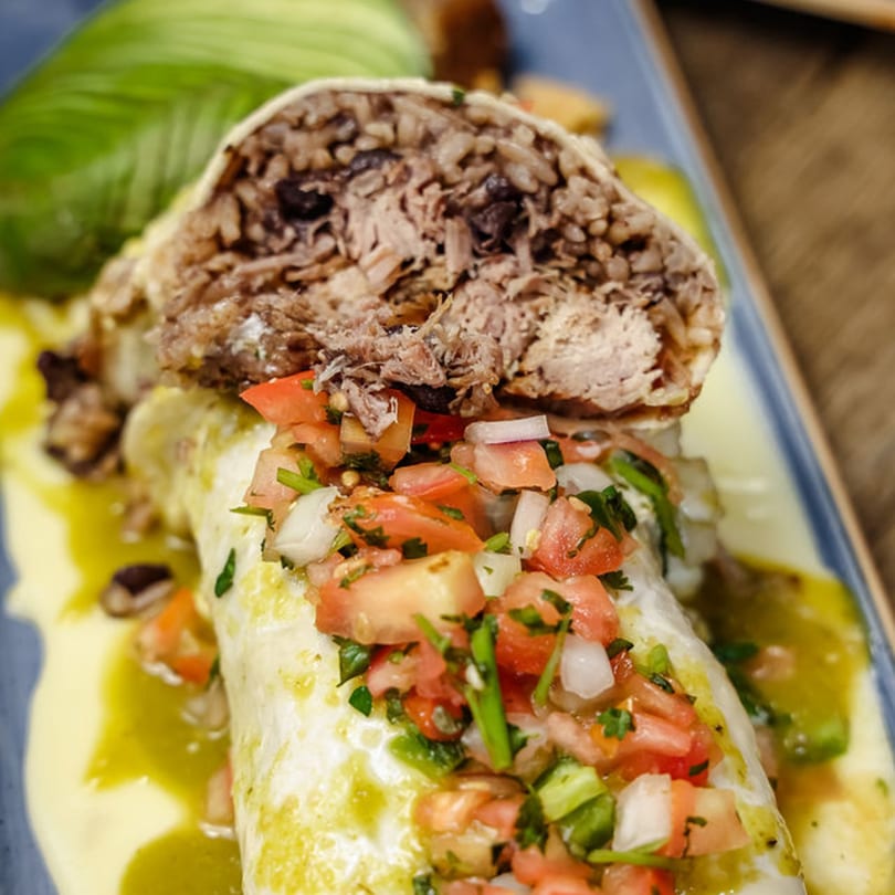 A large burrito, cut open to show shredded meat and rice, topped with pico de gallo and green sauce.