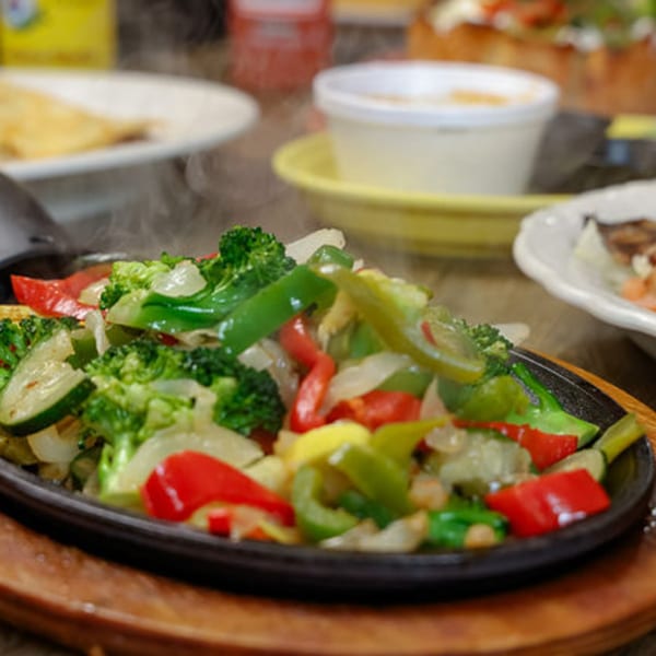 Steaming fajita vegetables, including broccoli, bell peppers, onions, and zucchini, on a sizzling cast iron plate.