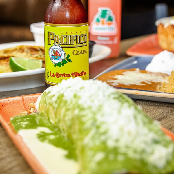 A Pacifico beer bottle stands next to a green-sauced burrito and other Mexican dishes.