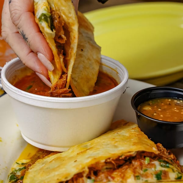 A hand dips a crispy birria taco into a white cup of consommé, with another taco and salsa nearby.