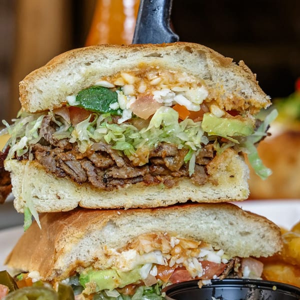 A large torta sandwich, cut in half, revealing steak, lettuce, avocado, tomato, and cheese.