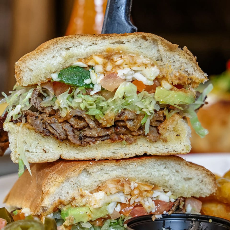 A large torta sandwich, cut in half, revealing steak, lettuce, avocado, tomato, and cheese.
