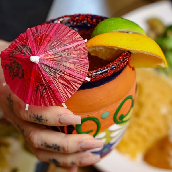 Hand holding a colorful, decorated clay mug with a drink, garnished with citrus and a red umbrella.