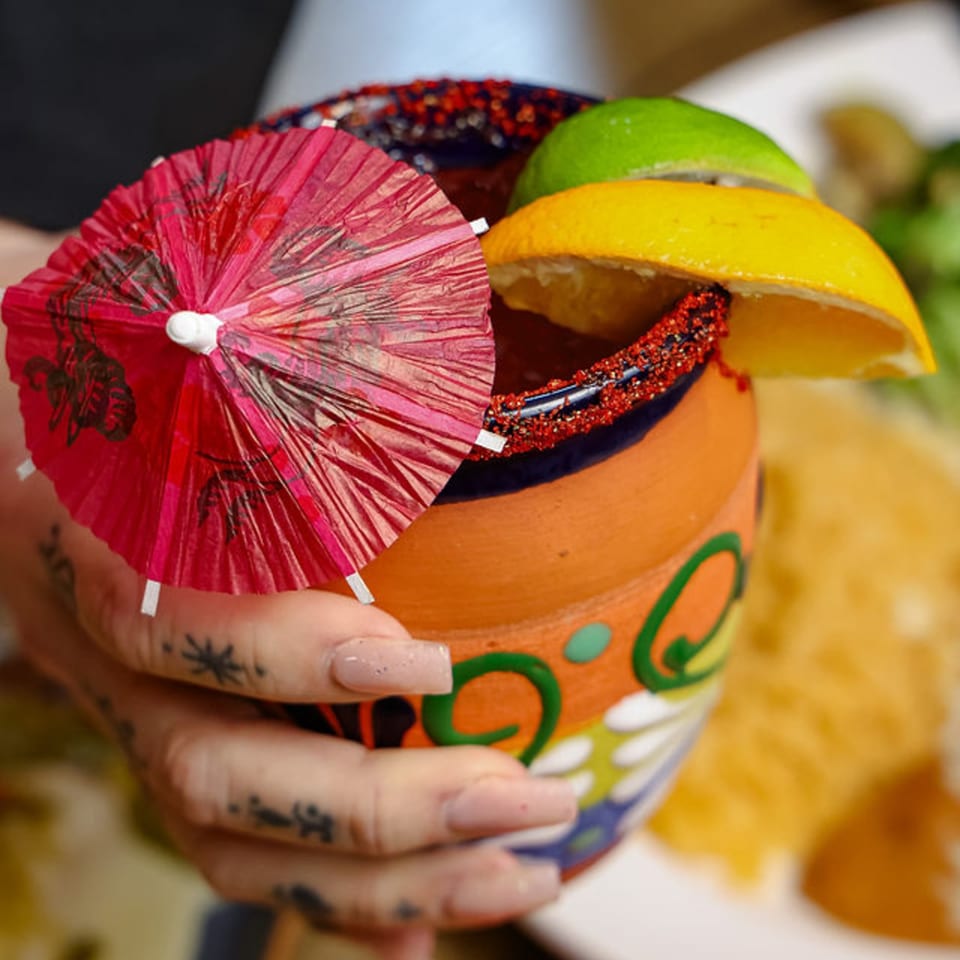 Hand holding a colorful, decorated clay mug with a drink, garnished with citrus and a red umbrella.